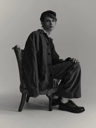 Monochrome portrait of a young adult male model wearing oversized workwear, seated on a distressed antique chair against a neutral backdrop.