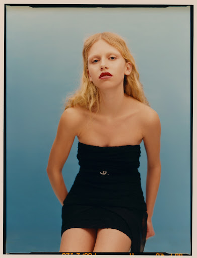 A blonde woman in a black strapless dress poses against a blue background, creating a fashion portrait with a clean and modern aesthetic.