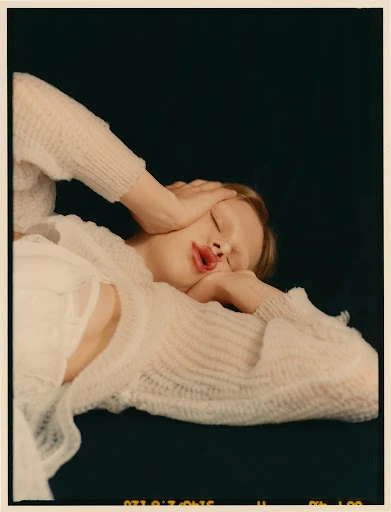 A young adult woman is lying down with her hands on her head, wearing a knitted top, against a solid dark background in a studio-style setting.
