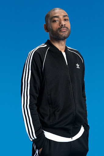 Portrait of Black man in Adidas track suit, looking directly at the camera against a bright blue studio backdrop.