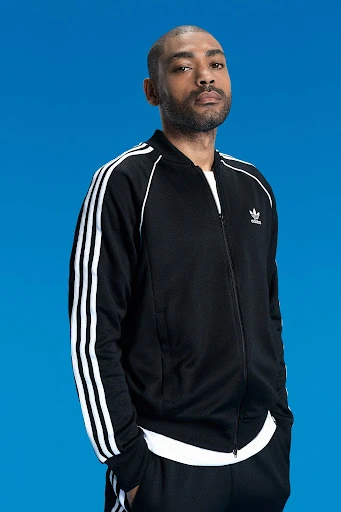 Portrait of Black man in Adidas track suit, looking directly at the camera against a bright blue studio backdrop.