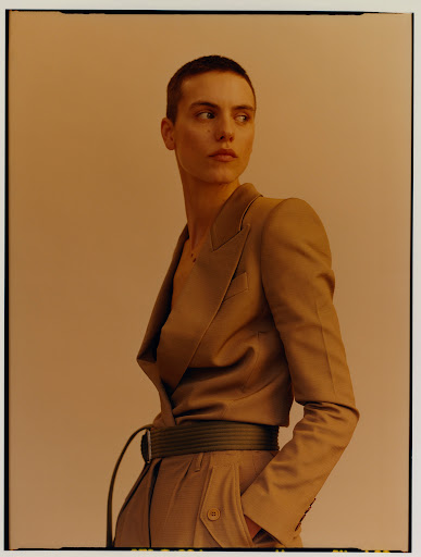 A person with a buzzcut wears a beige suit and belt and looks to the side in a studio with a light peach-colored backdrop.