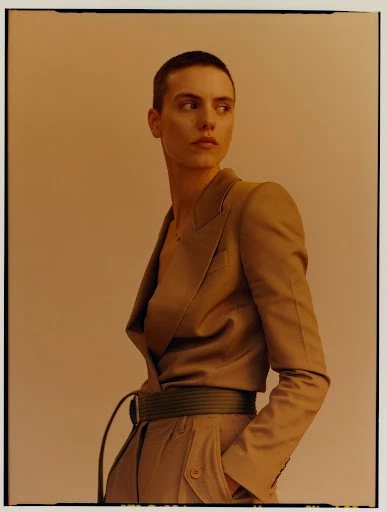 A person with a buzzcut wears a beige suit and belt and looks to the side in a studio with a light peach-colored backdrop.