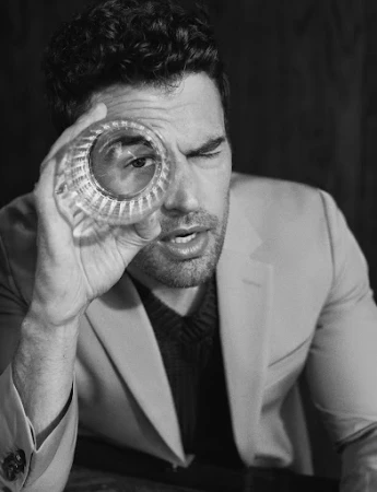 Monochrome portrait of a man holding a faceted glass up to one eye, wearing a light jacket over a dark ribbed sweater.