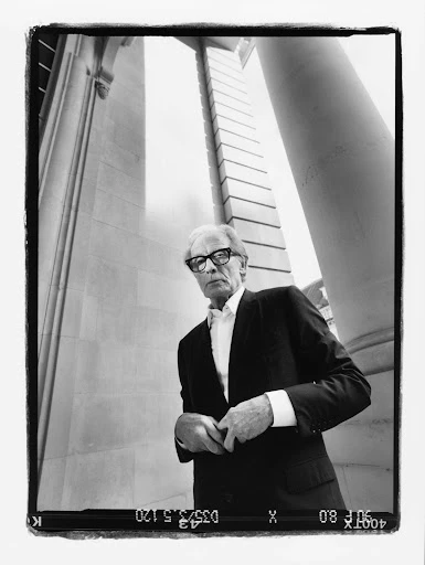 Monochrome portrait of an adult man in a suit and glasses stands outside a stone building, shot from a low angle with a wide lens.