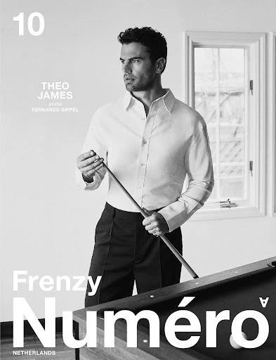 Black and white studio shot of Theo James holding a pool cue in a formal shirt and pants next to a pool table and a window.