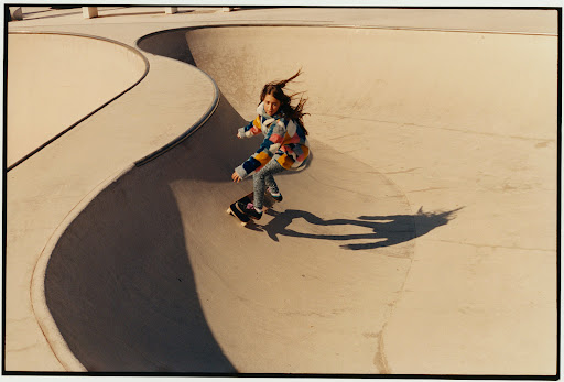 Young girl skateboarding on a concrete ramp in a skatepark, wearing colorful jacket and leggings, with dynamic pose and shadow.