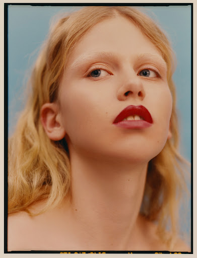 Close-up portrait of a young woman with light blonde wavy hair, fair skin, and red lipstick against a soft blue background.