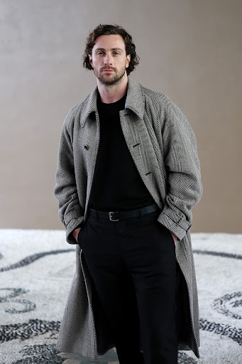Portrait of adult male with curly dark hair wearing a long grey herringbone coat and black shirt against a neutral background.