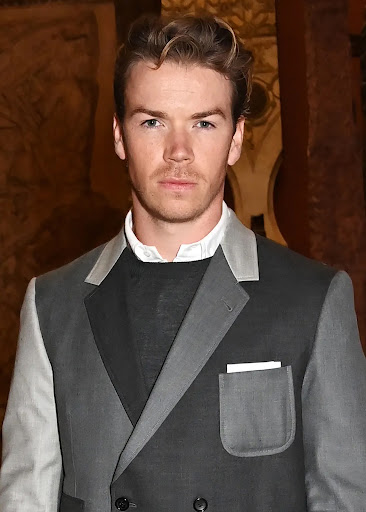 Portrait of Will Poulter in a grey two-tone suit and black sweater against a patterned background. He looks directly at the camera.