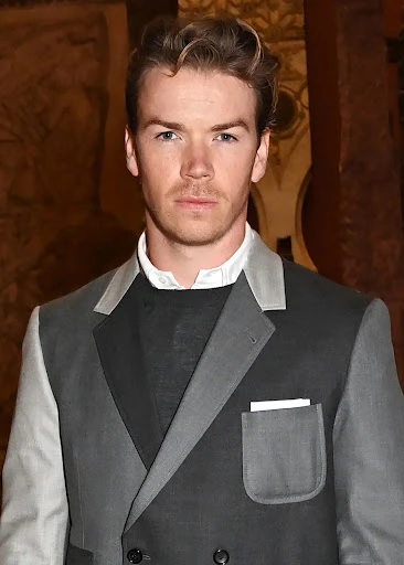 Portrait of Will Poulter in a grey two-tone suit and black sweater against a patterned background. He looks directly at the camera.
