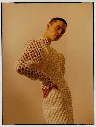 Portrait of a person with closely cropped hair wearing an elaborate crocheted dress while posing in front of a neutral background.
