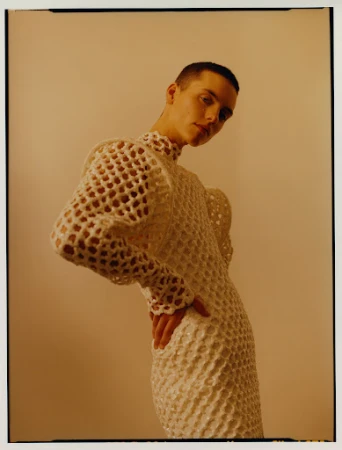 Portrait of a person with closely cropped hair wearing an elaborate crocheted dress while posing in front of a neutral background.