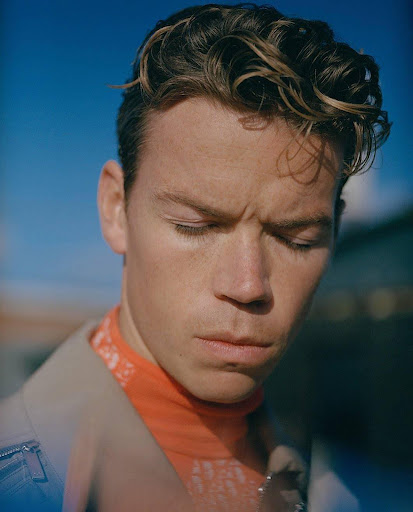 Close-up portrait of a man with curly brown hair wearing an orange turtleneck under a jacket against a clear blue sky.