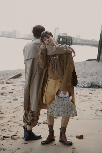 Two people standing on a sandy beach by the water holding a woven handbag while wearing layered tan trench coats and boots.