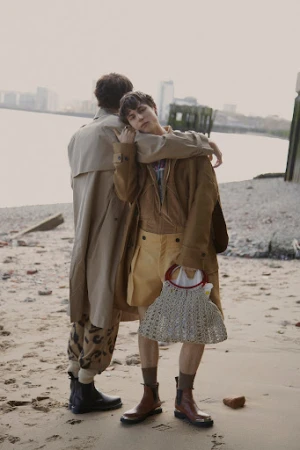 Two people standing on a sandy beach by the water holding a woven handbag while wearing layered tan trench coats and boots.