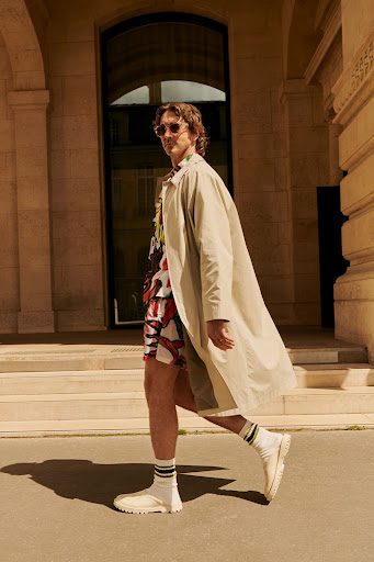 Man in sunglasses and coat walking past building in city on sunny day. Colorful patterned shirt and socks add a touch of style.