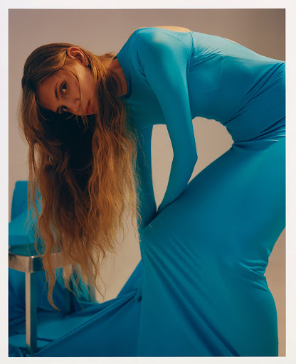 A woman with long brown hair and a turquoise dress poses next to a matching turquoise chair, creating a stylish fashion portrait.
