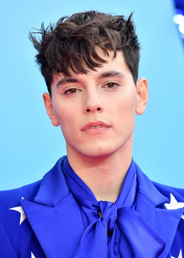 Close-up of a young adult male with dark hair, wearing a blue suit with white stars, standing against a bright blue backdrop.