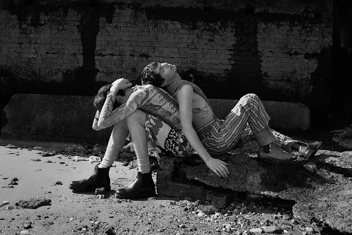 Monochrome fashion editorial image of two young men seated back-to-back on rocks near a weathered brick wall and sandy ground.
