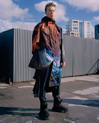 An adult male stands on concrete in a colorful hooded jacket and dark pants with a corrugated metal fence and building in the background.