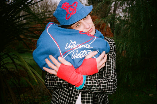 Young adult posing outdoors in bucket hat and patterned jacket, holding a blue sweatshirt with white script logo in a medium shot.