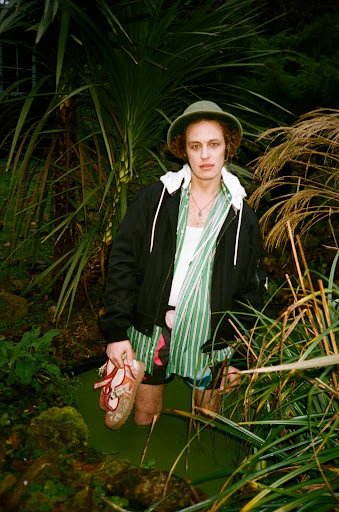 Man stands waist-deep in water in a garden, wearing a hat and jacket, holding shoes, surrounded by tropical greenery and foliage.