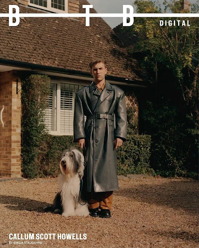 Man in grey trench coat poses with a bearded collie dog in front of a brick house with green trees. Fashion editorial.