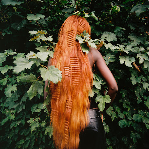 Woman with long, crimped auburn hair, leather pants with studded belt, and dark skin tone posing outdoors amongst green leafy shrubs.