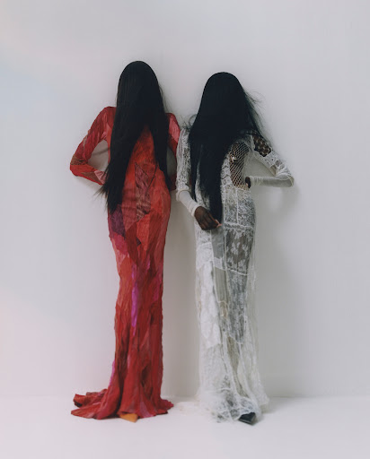 Two tall, slender fashion models with black hair covering their faces wear long, ornate gowns against a neutral backdrop in a studio setting.