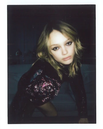 Polaroid photograph of a woman with blonde hair and dark eyeliner leaning forward in a dimly lit room