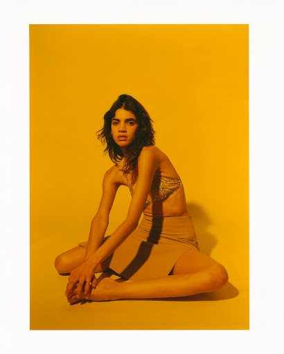 Fashion shot of a female model sitting on a yellow background wearing a top and skirt, posing with her arms resting on her knees.