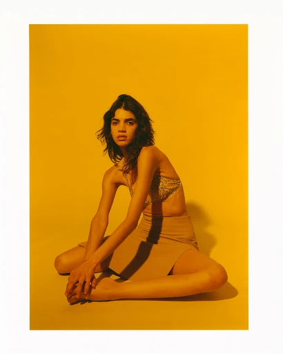 Fashion shot of a female model sitting on a yellow background wearing a top and skirt, posing with her arms resting on her knees.