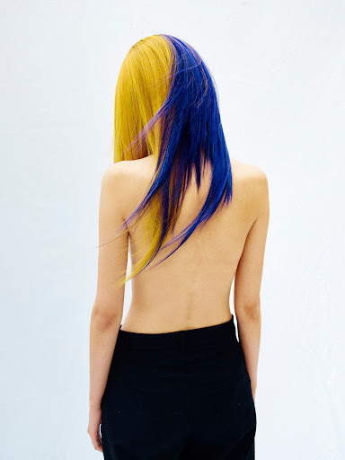 Back view of a slender woman with half yellow, half blue dyed hair, wearing black pants against a bright white background in a studio.