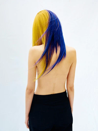 Back view of a slender woman with half yellow, half blue dyed hair, wearing black pants against a bright white background in a studio.