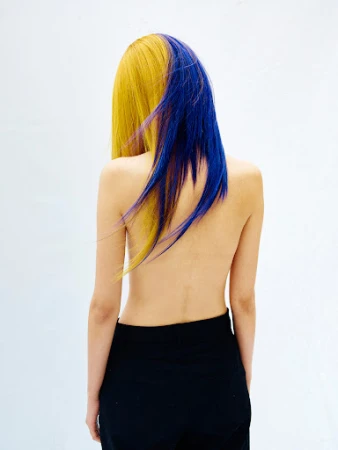Back view of a slender woman with half yellow, half blue dyed hair, wearing black pants against a bright white background in a studio.