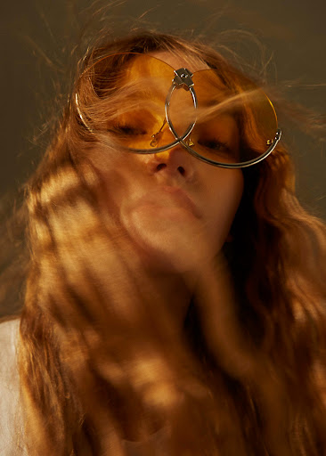 Portrait of a blonde woman wearing yellow sunglasses and a white shirt, with soft diffused lighting creating a hazy effect.