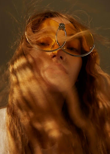 Portrait of a blonde woman wearing yellow sunglasses and a white shirt, with soft diffused lighting creating a hazy effect.