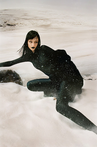 A woman in a black bodysuit kneels in snow against a stark, wintry backdrop with rocks and a distant snowy horizon.