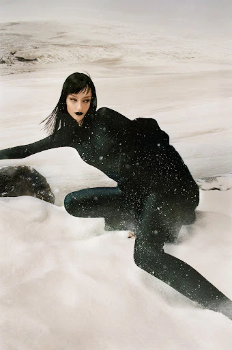 A woman in a black bodysuit kneels in snow against a stark, wintry backdrop with rocks and a distant snowy horizon.