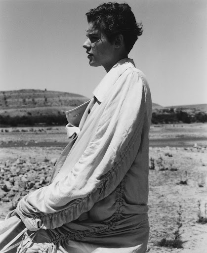 Side profile of a young adult in an outdoor setting, wearing a light-colored coat, with a background of dry, hilly landscape and clear sky.