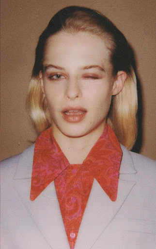 Close-up of a blonde adult female winking and wearing a pastel jacket with a large pink patterned collar, looking directly at the camera.