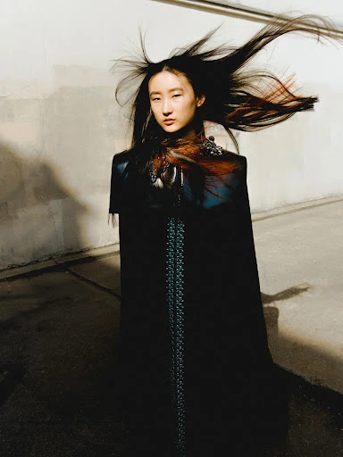 Asian woman in long black coat poses with wind-blown dark hair and feathered shoulder adornments against a light-grey textured wall.