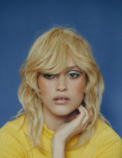 Close-up portrait of a blonde adult woman with light skin wearing a yellow top against a blue background in a studio setting.