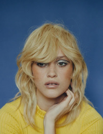 Close-up portrait of a blonde adult woman with light skin wearing a yellow top against a blue background in a studio setting.