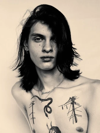 Close-up portrait of a young man with long dark hair, chest tattoos, and layered necklaces against a light background.