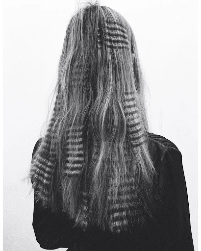 Back view of a woman with long, gray, wavy hair styled with patterned hair clips, set against a plain white studio backdrop.