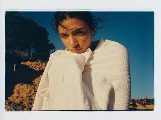Portrait of a young woman wrapped in a white textured robe against a blue sky and rocky landscape. Natural light, serene mood.