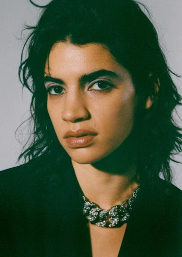 Close-up studio shot of a young adult female model with dark hair wearing a black blazer and chunky rhinestone necklace, posing against a grey backdrop.