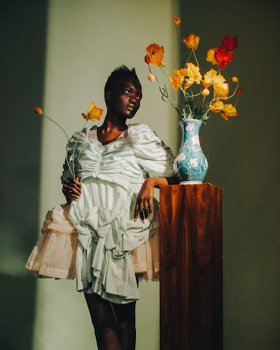 Portrait of a Black woman in a white dress with floral accents, posing next to a wooden pedestal with a vase of orange flowers.